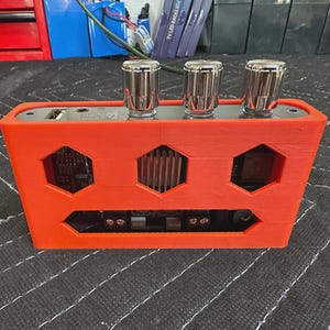 以下が含まれることがあります： 上部に3つの銀色のノブが付いたオレンジ色の電子デバイス。デバイスは、六角形の切り欠きと下部の長方形の開口部を備えた長方形の形状をしています。デバイスは黒い表面にあります。