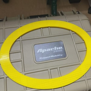 Puede incluir: Una junta circular amarilla y texturizada descansa sobre una maleta beige. La maleta tiene el texto "Apache 2800 Rugged Mobility" en una placa plateada. El color brillante de la junta contrasta con la maleta.