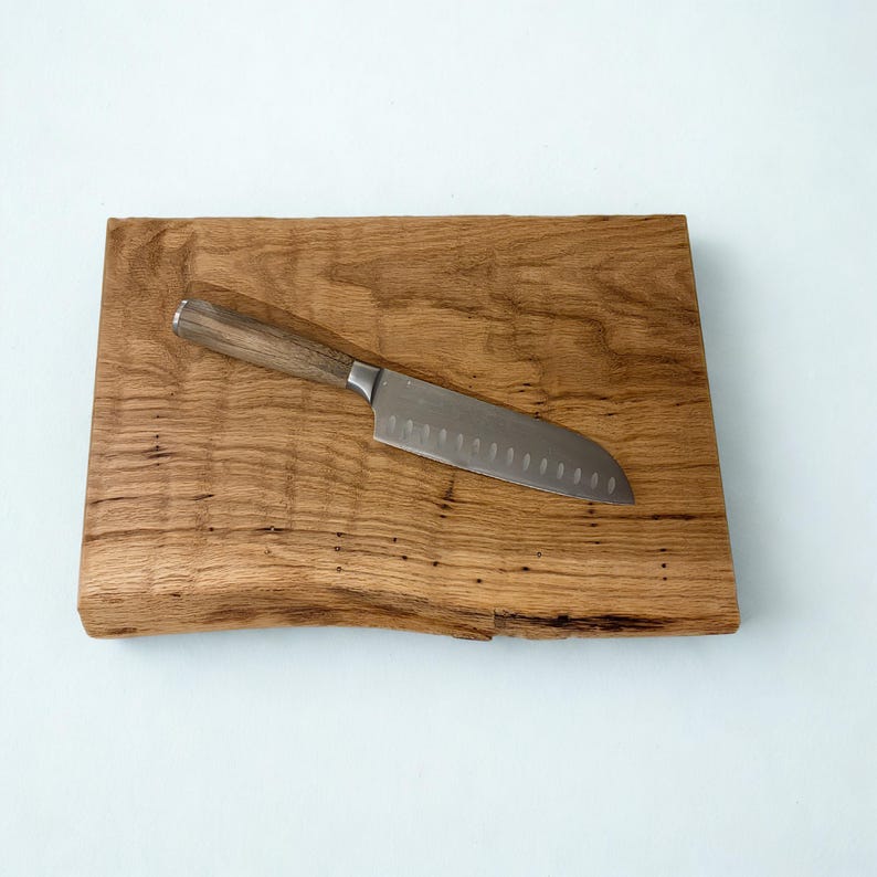 May include: A wooden cutting board with a live edge, featuring a stainless steel and wood-handled chef's knife. The board is made of light brown wood with a natural grain pattern. The knife is approximately 12 inches long.