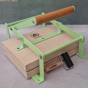 May include: A light green metal book press with a wooden handle and two light-colored wooden boards. The press features the InkuMori logo on the top board. A small black rubber brayer with a wooden handle and the InkuMori logo is placed on top.