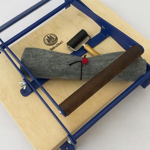 Pode incluir: Uma prensa de impressão azul com base de madeira, segurando um cobertor de feltro cinza, um rolo pequeno e um rolo de madeira escura. A base de madeira tem o texto "InkuMori" e um logotipo.