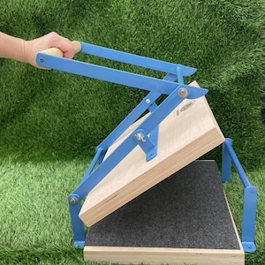 Puede incluir: Una prensa de encuadernación de metal azul y madera clara con un mecanismo de palanca y un mango de madera. El dispositivo consta de dos tablas de madera gruesas, con una almohadilla de fieltro gris oscuro unida a la tabla inferior, conectadas por brazos de metal azul.