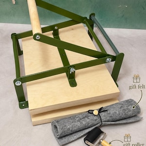 Puede incluir: Una prensa de encuadernación con base y placa superior de madera clara, marco de metal verde oliva oscuro y mango de madera. Incluye un fieltro gris enrollado con la etiqueta gift felt y un pequeño rodillo negro con la etiqueta gift roller.