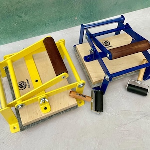 May include: Two printmaking presses, one yellow and one blue, with wooden handles and bases. Each press has a wooden roller and a small black ink roller. The presses are on a light gray surface.