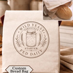 May include: A beige fabric bread bag featuring a brown circular graphic of a mason jar with the text Wild Yeast, Samantha's Starter, Fed Daily. The bag has dark brown side panels and a small label at the bottom reading Custom Bread Bag.