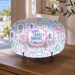 May include: An oval ceramic serving platter featuring a colorful pattern of Mahjong tiles with peacocks, flowers, and dragons. A central pink-bordered label displays the text BAMS, CRAKS & SNACKS at Olivia's in blue and pink lettering.