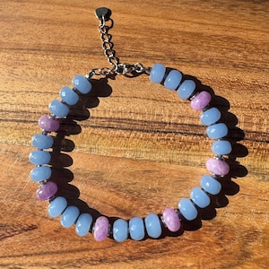May include: A beaded bracelet featuring alternating light blue and purple beads. The bracelet is strung on a silver chain with an adjustable clasp. The beads have a faceted, oval shape. The bracelet is displayed on a wooden surface.