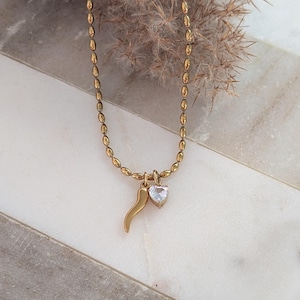 Puede incluir: Collar dorado con cadena de cuentas y dos colgantes. Un colgante es una pequeña piedra transparente en forma de corazón, y el otro es una cornicello dorado. El collar se muestra sobre una superficie de mármol blanco y gris.