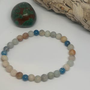 Puede incluir: Una pulsera de cuentas redondas en tonos azul claro, melocotón y blanco. También se ven una piedra verde y marrón y un manojo de salvia seca. La pulsera es probablemente para fines de moda o espirituales.