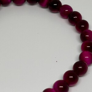 May include: A close-up of a bracelet made of round, polished beads. The beads are a deep magenta color with a subtle sheen. The bracelet is set against a plain white background, highlighting the jewelry.