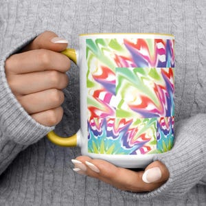 May include: White ceramic mug with a yellow handle and rim, featuring a vibrant, abstract design in shades of red, green, blue, and purple. The mug is held by a person wearing a gray sweater.
