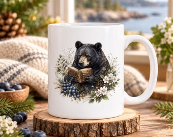 May include: A white ceramic mug featuring a detailed illustration of a black bear holding an open wooden book, surrounded by clusters of dark blue berries and small white flowers. The mug has a smooth finish and a standard handle on the right side.