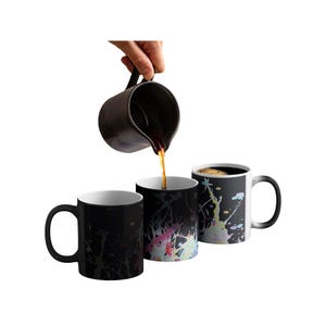 May include: Three black heat-sensitive mugs with white interiors. One mug is being filled with a dark liquid from a black pitcher. The mugs' designs change color when hot liquid is added, revealing colorful patterns.