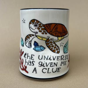 May include: White ceramic tumbler with a black rim, featuring a cartoon sea turtle with a brown shell and yellow accents. The tumbler has the text "the UNIVERSE has given ME A CLUE" and illustrations of crystals and coral.