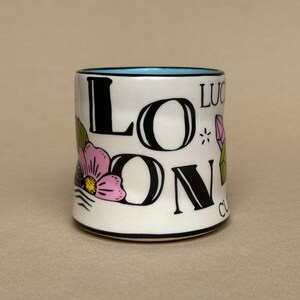 May include: A white ceramic mug with the word "LONDON" in large black letters, with a pink flower and green leaves. The mug has a blue interior and black trim. The word "LUCKY" is also printed on the mug.