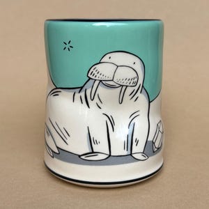 May include: A ceramic mug featuring a cartoon walrus design. The walrus is white with black outlines and tusks, set against a teal background with a star. The mug has a black rim and base, with a gray band near the bottom.
