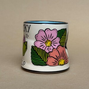 May include: A white ceramic mug with a blue interior rim, featuring pink and coral flowers with yellow centres and green leaves. The mug has the words "LUCKY" and "UP" printed on it.