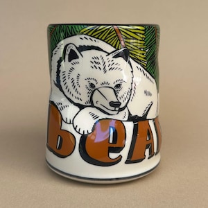 May include: A ceramic mug featuring a white bear illustration with the word "BEAR" in brown letters. The background includes green and yellow foliage. The mug has a glossy finish.