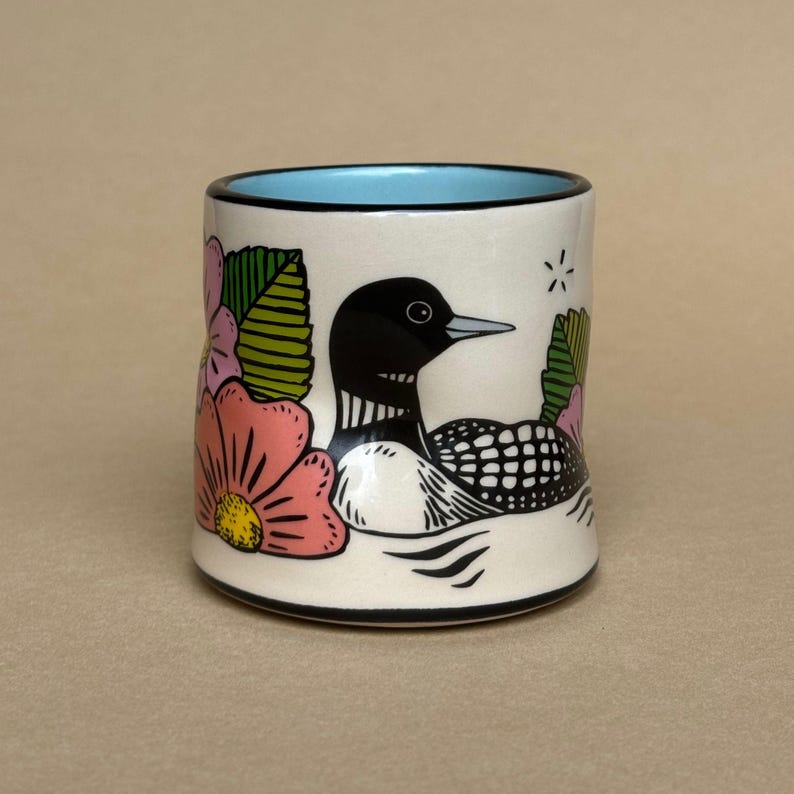 May include: A ceramic mug with a white base, featuring a black and white loon design, pink flowers, and green leaves. The rim is black, and the interior is light blue. A star graphic is also present.