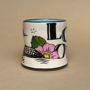May include: A white ceramic mug with a black rim and a light blue interior. The mug features a floral design with a pink flower, green leaves, and the word "LOON" in black. A black and white bird is also depicted.