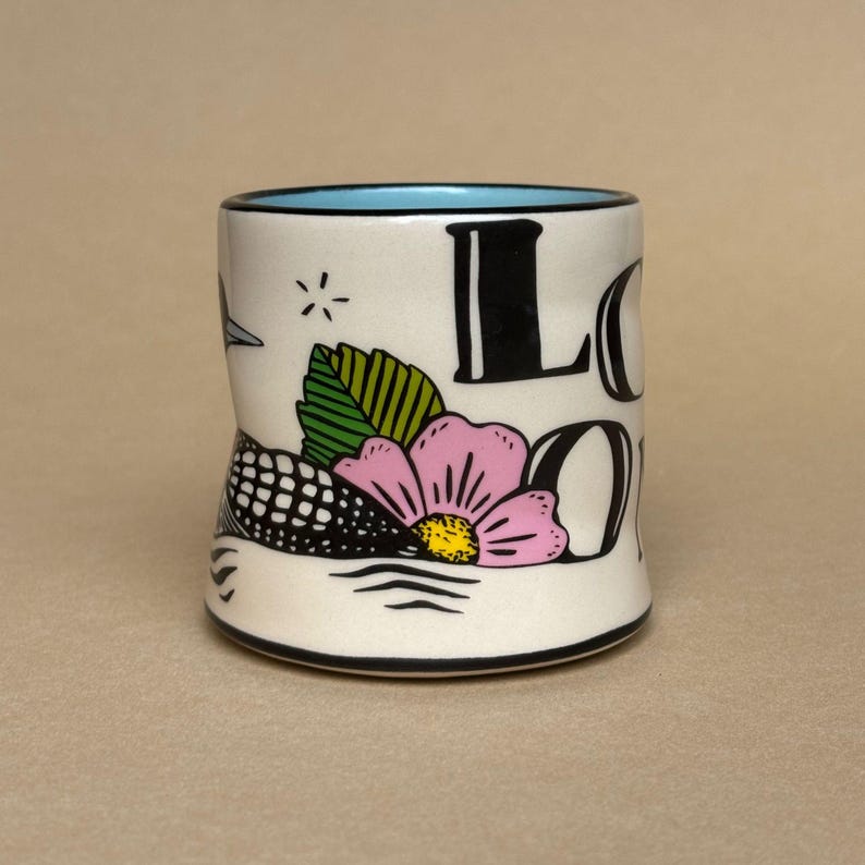 May include: A white ceramic mug with a black rim and a light blue interior. The mug features a floral design with a pink flower, green leaves, and the word "LOON" in black. A black and white bird is also depicted.