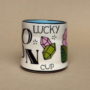 May include: A white ceramic cup with a black rim and a light blue interior. The cup features the words "ON LUCKY CUP" in black lettering, along with illustrations of pink and green crystals, a pink flower, and green leaves.