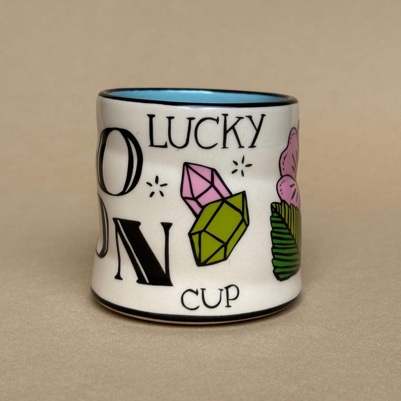May include: A white ceramic cup with a black rim and a light blue interior. The cup features the words "ON LUCKY CUP" in black lettering, along with illustrations of pink and green crystals, a pink flower, and green leaves.