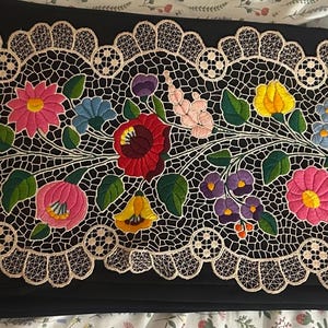 May include: A rectangular black fabric runner featuring a central panel of intricate cream-colored lace. The lace is decorated with vibrant, multi-colored floral embroidery in shades of red, pink, yellow, blue, and purple with green leaves.