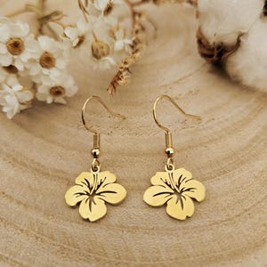 Peut inclure: Une paire de boucles d'oreilles en forme de fleur d'hibiscus dorée. Les boucles d'oreilles ont un design à crochet et sont présentées sur une surface en bois avec des fleurs blanches et du coton en arrière-plan. Bijoux floraux.