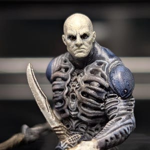 May include: A detailed figurine of a bald, pale-skinned humanoid figure with a stern expression. The figure is clad in armor with intricate, textured designs in shades of gray and blue. It holds a silver blade, suggesting a fantasy or sci-fi theme.