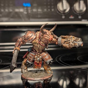 May include: A detailed miniature figurine of a minotaur warrior. The figure is painted with a combination of brown, red, and tan hues, with metallic accents. It is posed with a sword and a large, ornate gauntlet. The minotaur stands on a circular base.