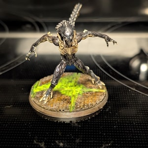 May include: A detailed figurine of an Alien Xenomorph, posed with outstretched arms. The figure is primarily metallic gray with yellow accents on the chest and head. It stands on a circular base with a grid pattern and green and brown coloring.
