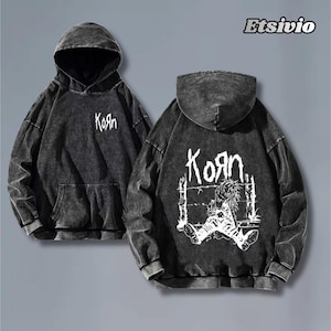 May include: Two charcoal grey hoodies featuring the word "Korn" in white. One hoodie displays "Korn" on the front. The other has a graphic and "Korn" on the back. The hoodies have a washed effect.