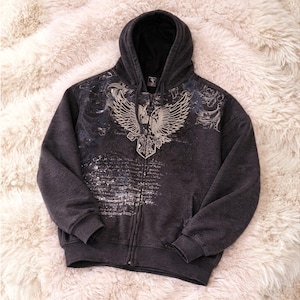 Könnte beinhalten: Anthrazitfarbener Kapuzenpullover mit Reißverschluss und schwarzer Kapuze. Auf der Vorderseite befindet sich eine detaillierte silber- und graue Grafik eines Adlers mit ausgebreiteten Flügeln. Das Design umfasst Schrift- und Dekorationselemente.