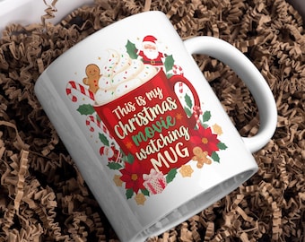 Może przedstawiać: Biały ceramiczny kubek z czerwonym wzorem z napisem "This is my Christmas movie watching MUG". Kubek zawiera ilustracje Świętego Mikołaja, piernikowych ludzików, lasek cukrowych i gwiazd betlejemskich. Kubek ma biały uchwyt.