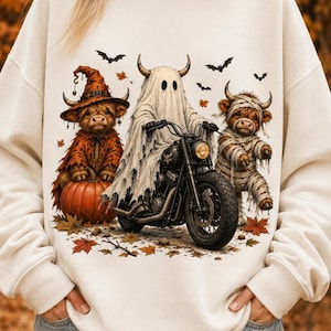 Op de afbeelding: Een crèmekleurig sweatshirt met een grafische print van een geest op een zwarte motorfiets, geflankeerd door een Highland-koe met een heksenhoed naast een pompoen en een in mummiewindsel gewikkelde Highland-koe, met vleermuizen en herfstbladeren.