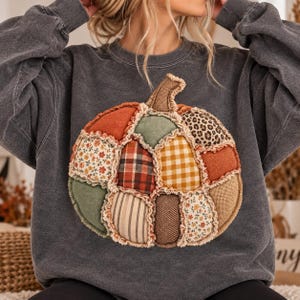 Op de afbeelding: Een antracietgrijze sweater met ronde hals en een grote, centrale pompoenafbeelding bestaande uit diverse stoffen lapjes. Het ontwerp bevat secties met ruitpatronen, gingham, bloemenprints, luipaardprint, jute en effen gekleurde stoffen.