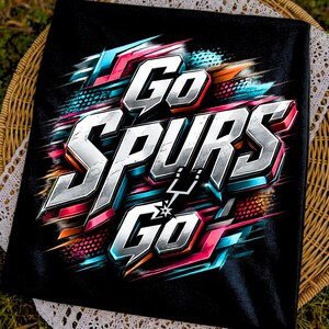 May include: A black t-shirt featuring a vibrant graphic design with the text Go SPURS Go in a bold, metallic, three-dimensional font. The lettering is surrounded by dynamic streaks of neon pink, cyan, and orange, with a small star icon included.