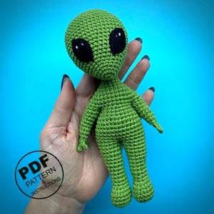 May include: A green crocheted alien doll with large black eyes. The doll is handmade and has a detailed, textured surface. The image includes a circular graphic that says "PDF PATTERN & INSTRUCTIONS".