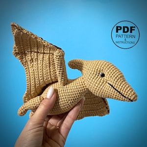 May include: A tan crocheted Pterodactyl toy with black accents, held in a hand. The wings are spread, and the head has a small horn. The text "PDF PATTERN & INSTRUCTIONS" is in a circle in the upper right corner.