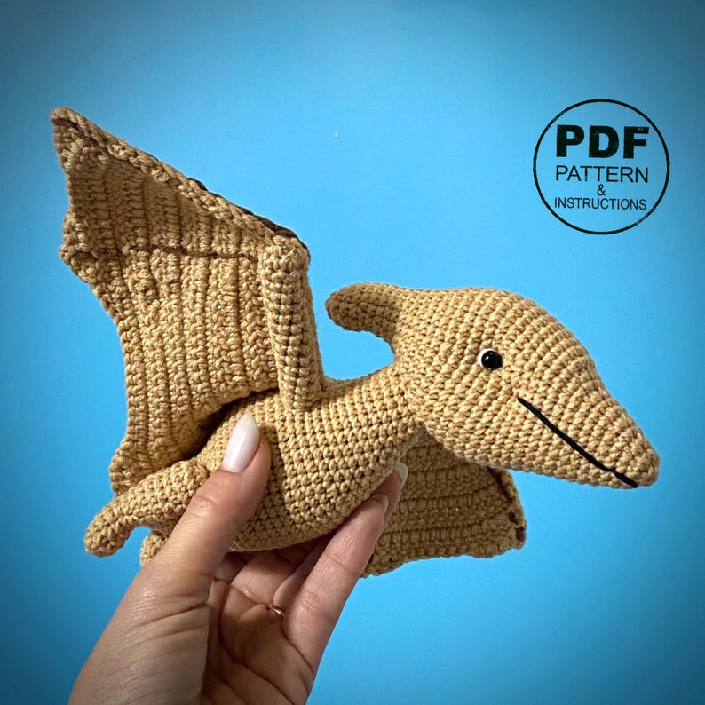 May include: A tan crocheted Pterodactyl toy with black accents, held in a hand. The wings are spread, and the head has a small horn. The text "PDF PATTERN & INSTRUCTIONS" is in a circle in the upper right corner.