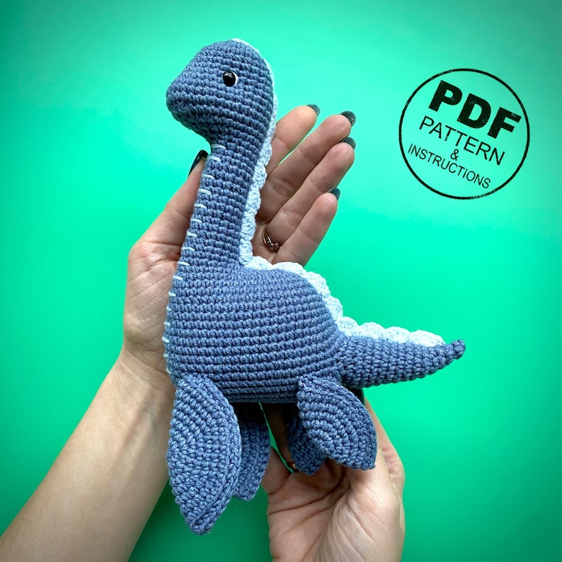 May include: A handmade blue crochet dinosaur toy with a light blue belly and accents. The toy has small black eyes and is held in a person's hands. A circular graphic with the text "PDF PATTERN & INSTRUCTIONS" is in the background.