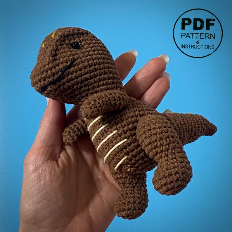 May include: A brown crocheted dinosaur toy with white stripes on its side. The dinosaur has a smiling mouth and small yellow accents on its head. The image includes the text "PDF Pattern & Instructions" in a circular design.
