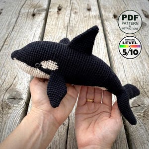 May include: A crocheted orca whale in black and white, held in hands. The whale has a dorsal fin, pectoral fins, and a tail fin. The image includes a PDF pattern and instructions, with a difficulty level of 5/10.