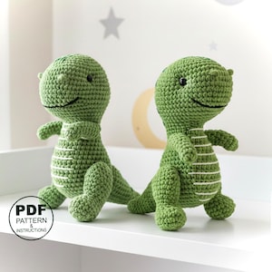 Puede incluir: Dos juguetes de dinosaurio verdes de ganchillo con rayas blancas en la barriga. Los dinosaurios tienen pequeños ojos negros y están sentados sobre una superficie blanca. La imagen incluye un gráfico circular con el texto "PDF PATTERN & INSTRUCTIONS".