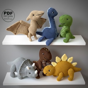Könnte beinhalten: Eine Sammlung von sechs handgefertigten gehäkelten Dinosaurierspielzeugen in verschiedenen Farben, darunter Beige, Blau, Grün, Grau, Braun und Gelb. Die Dinosaurier werden auf weißen Regalen vor einer grauen Wand ausgestellt. Der Text "PDF PATTERN & INSTRUCTIONS" befindet sich oben links.