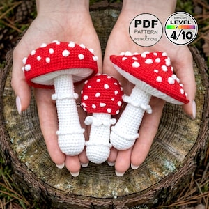 May include: Three crocheted mushroom figures with red caps and white spots are held in open hands. The stems are white with a ruffled design. The image includes text that says "PDF Pattern & Instructions" and "Difficulty Level 4/10".