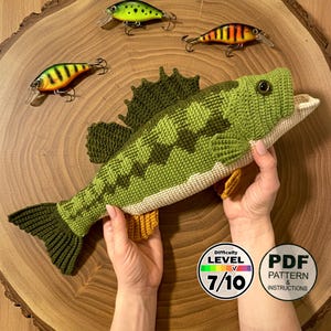 May include: A handmade crochet fish in shades of green, with a cream-colored belly and yellow fins. The fish is displayed with three fishing lures. A circular graphic indicates a difficulty level of 7/10 and a PDF pattern is available.