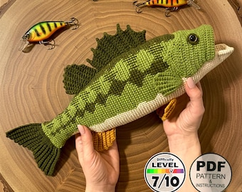 May include: A handmade crochet fish in shades of green, with a cream-colored belly and yellow fins. The fish is displayed with three fishing lures. A circular graphic indicates a difficulty level of 7/10 and a PDF pattern is available.