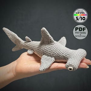 May include: A crocheted gray hammerhead shark toy. The shark has a distinctive hammer-shaped head, fins, and a tail. The image includes a circular graphic with the text "Difficulty Level 5/10" and another with "PDF Pattern & Instructions".